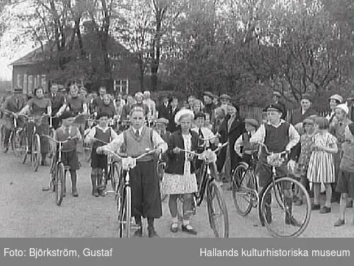 Cykelns Dag i Varberg, anordnad av Monark. I förgrunden syns några barn från Rosenfredsskolan. Flickan i vit basker heter Maj, gift Andersson. De hade cyklar som de lånat från Monark. Bakom dem syns fyra Monark-kontorister med nya cykelmodeller. Enligt fotografens liggare är motivet från "Sexdagarsloppet", ett amatörcykellopp som startades år 1924.