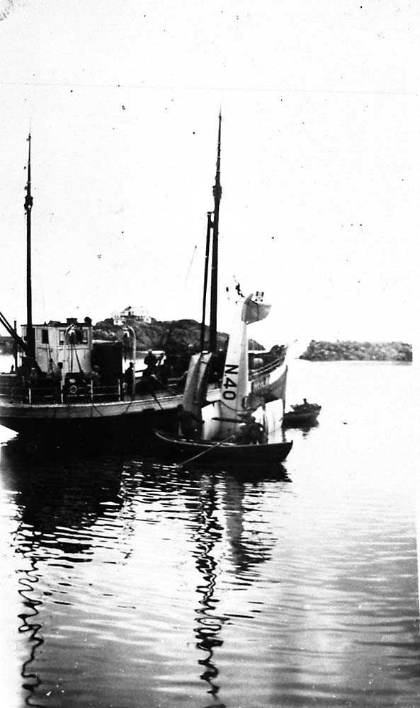 Ett sjøfly som blir heist ombord i en båt, etter et havari, Saaski "Måsen". To robåter på vannet. Flere personer ombord i båtene.