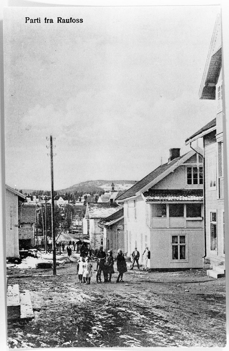 Avfotografert postkort fra Raufoss ca. 1915. Bildet er tatt nedover Østvollvegen mot torvet. Man kan se en baker stå utafor Andresens Bakeri. Jenta til høyre er Constanse Løkken.