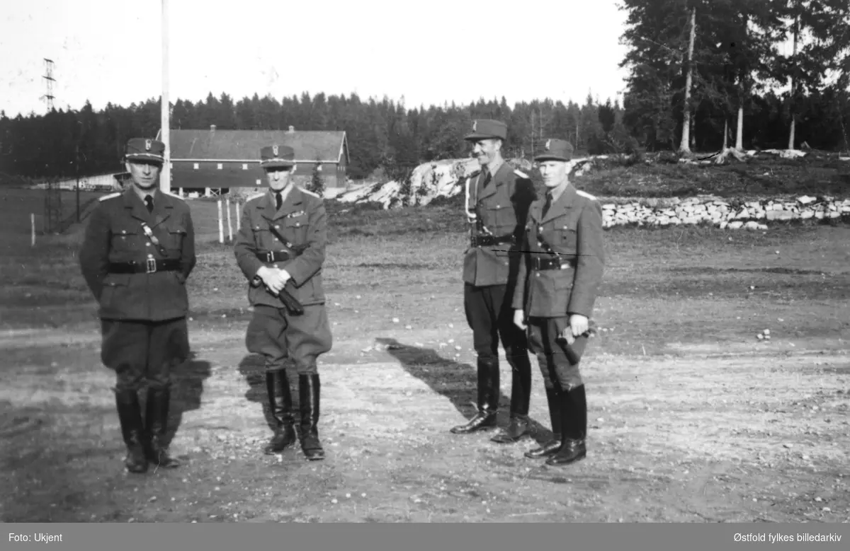 Arbeidstjenesten (AT-leir)  i Varteig ca. 1942-43. Høyere befal. Til høyre nest-sveitfører Krosby, nummer to fra venstre er distriktssjef Grahm.  Gårdsbruk i bakgrunnen, like ved Varteig kirke.