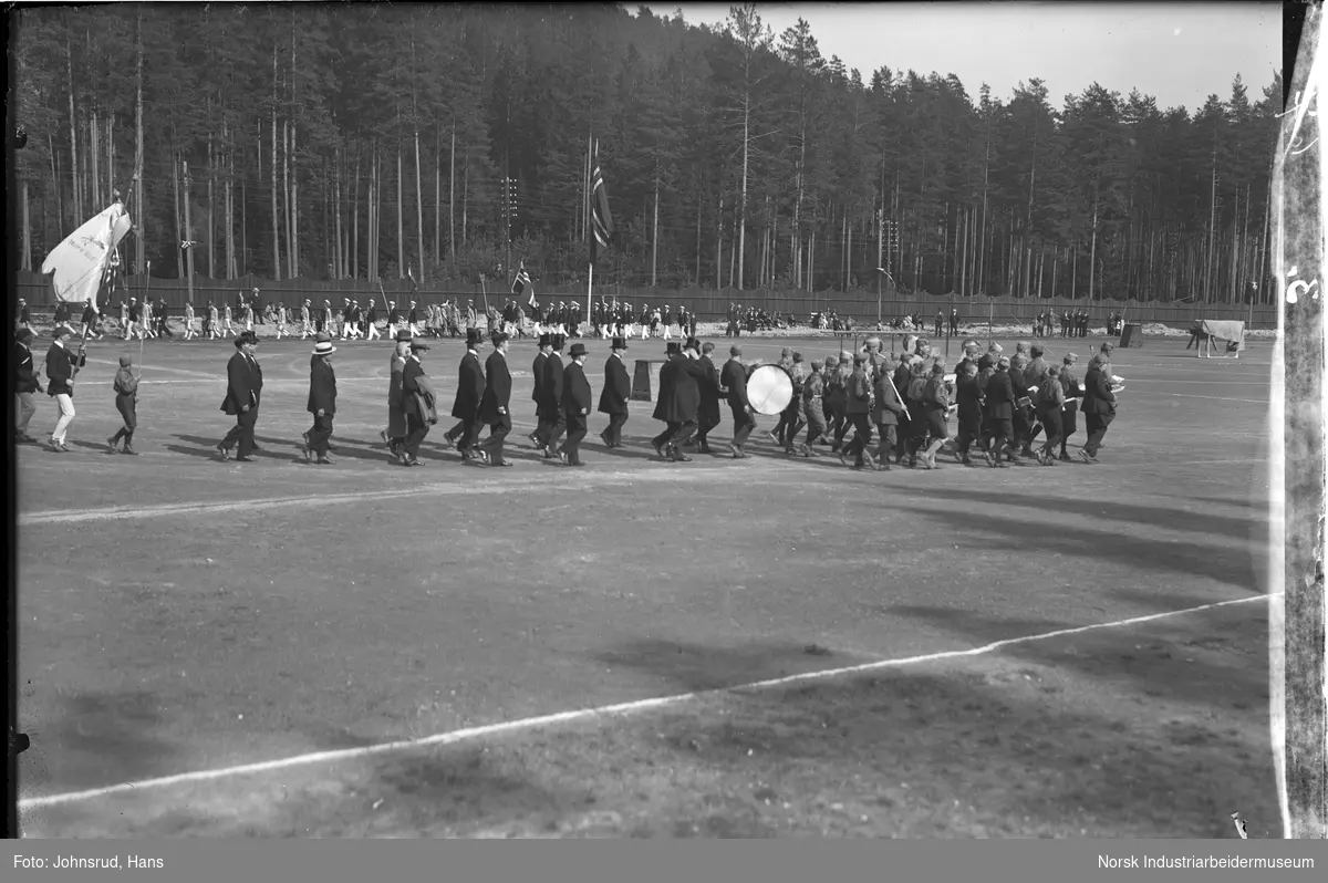 Korps marsjerende inn på idrettsbane under turnstevne. Utøvere gående i opptog bak.