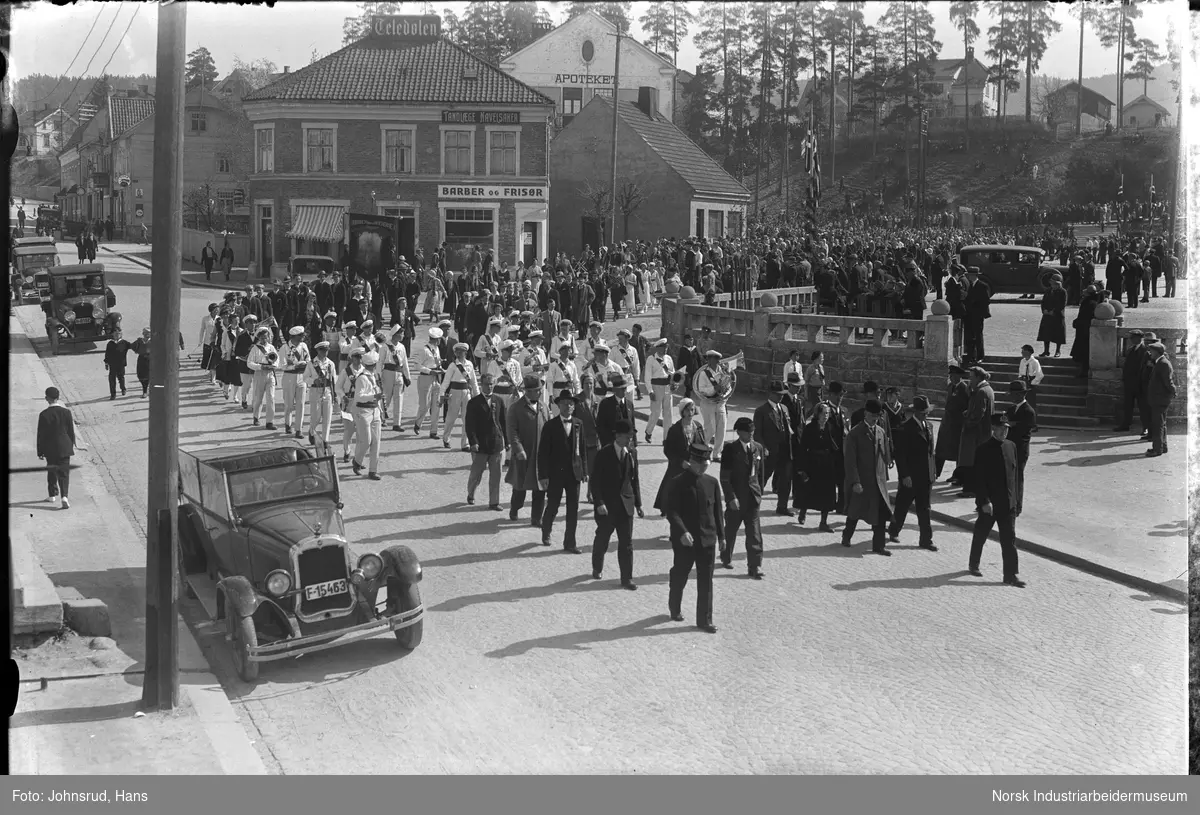 17. mai feiring 1932. Korps marsjerer forbi torget. Biler parkert langs gaten.