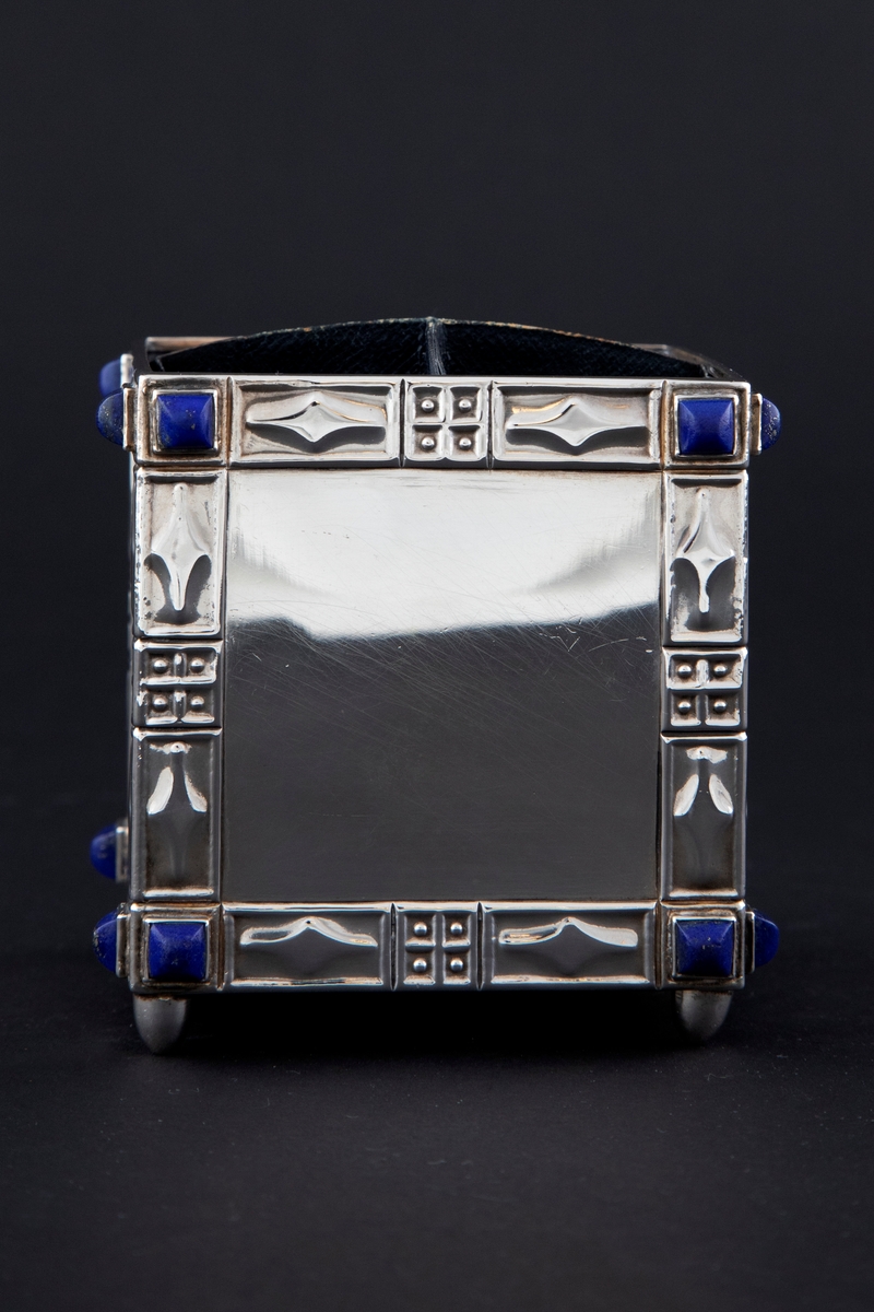 Kvadratisk formet sigarettboks på fire føtter. De fire sidene består av ornamentale rammer, hver med lapis lazuli (lasursteiner) i hjørnene. Det indre har en bevegelig korsformet inndelingsvegg i blått skinn festet i slisser.