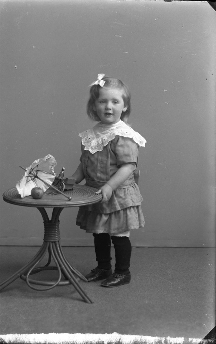 En liten flicka står vid ett lågt runt bord. På bordet ligger det ett litet parasoll, en leksaksbil och ett äpple.