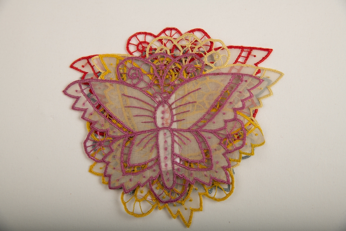 Brikke i form av en sommerfugl med utspilte vinger. Brodert i ulike farger med silketråd med knapphullsting og uttrekksøm.