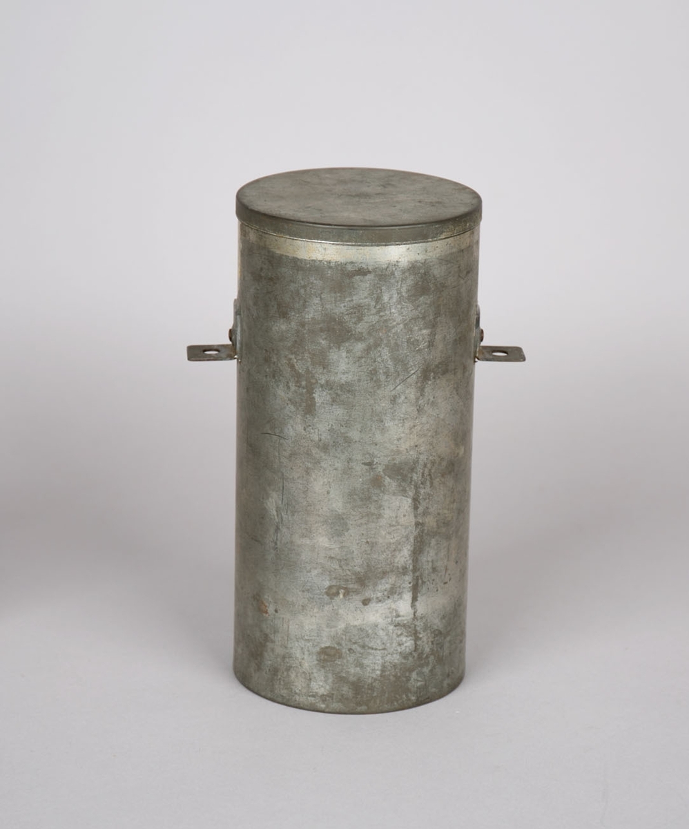 Metallbeholder med lokk. Del av luftvernskyts fra krigen 1939-45. Sylinderformet beholder med bunn i ene ende og løst lokk i andre ende. To vinkler/festebeslag montert på hver side av beholder. 