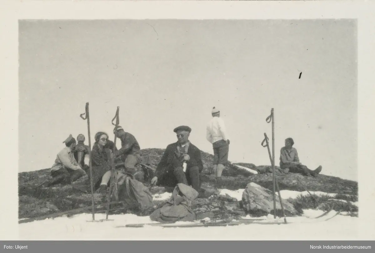 Påsken 1925 på Møsstrond. Mennesker iført friluftstøy sitter på en steinknaus og tar seg en pause fra skituren. Ski, ryggsekker og staver ligger i forkant.