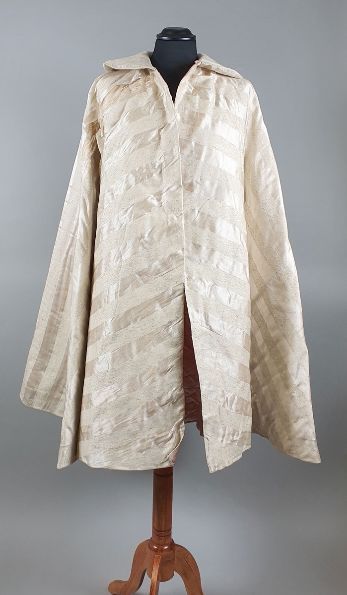 Hvit cape med diagonale striper vevd i lin og silke. Rosa fôr av bomull. Capen er vattert. To lommer på innsiden av hvert forstykke. Bred skjortekrage med buede kanter.