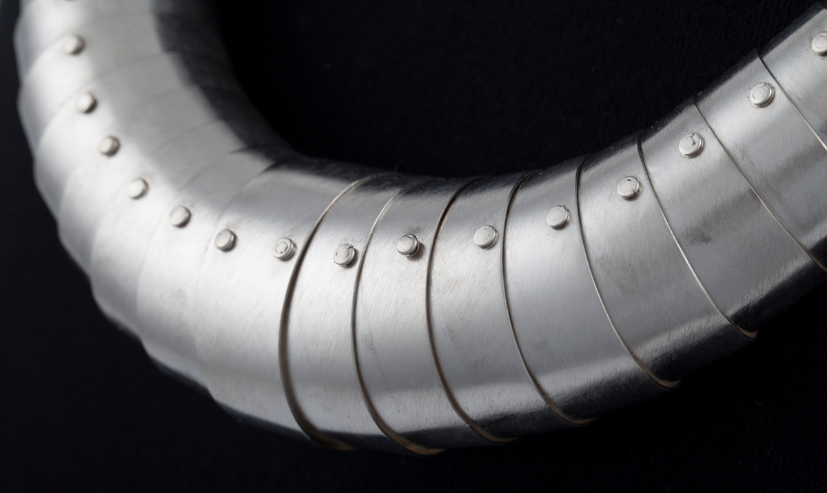 Slangeformet halssmykke i sølv bestående av 55 bevegelige ledd som er tredd i hverandre og minker i størrelse fra midt foran og mot låsen i nakken. Smykket har en symmetrisk form.