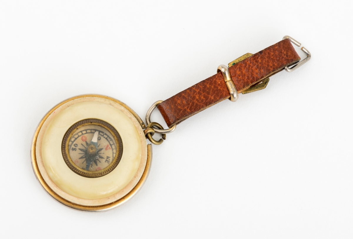 Et rundt kompass i lommeformat med reim. Selve kompasset er satt inn i skiver av antatt bakelitt. Reima er laget av lær. Beslag av messing.