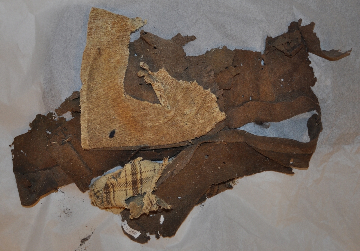 10 fragment av kavaj i brunt ylletyg som tillhört Fraenkels kavaj. I en av fickorna fanns en liten del av en blyertspenna.