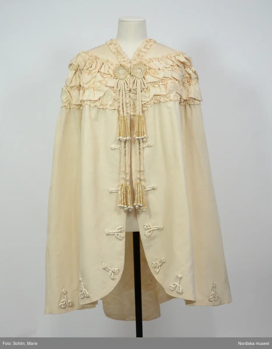Ordered by Ingrid Bergström née Anjou (1856–1948), married to cabinet minister David Bergström, a major supporter of the right to vote for all. Delivered 1906. White silk cape with decorative agraffs and tassels. Worn by Ingrid Bergström at the christening of 
Prince Gustaf Adolf in the Royal Chapel in 1906.