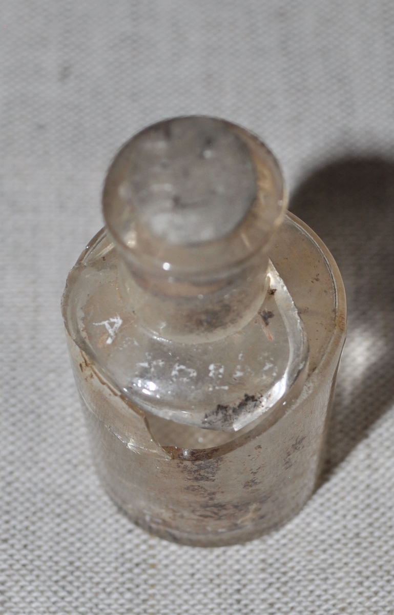 Cylindriskt fragment av glasflaska, tillhör flaskan (APXP.248) och var fastsatt vid denna. Har haft anilinfärgat innehåll.