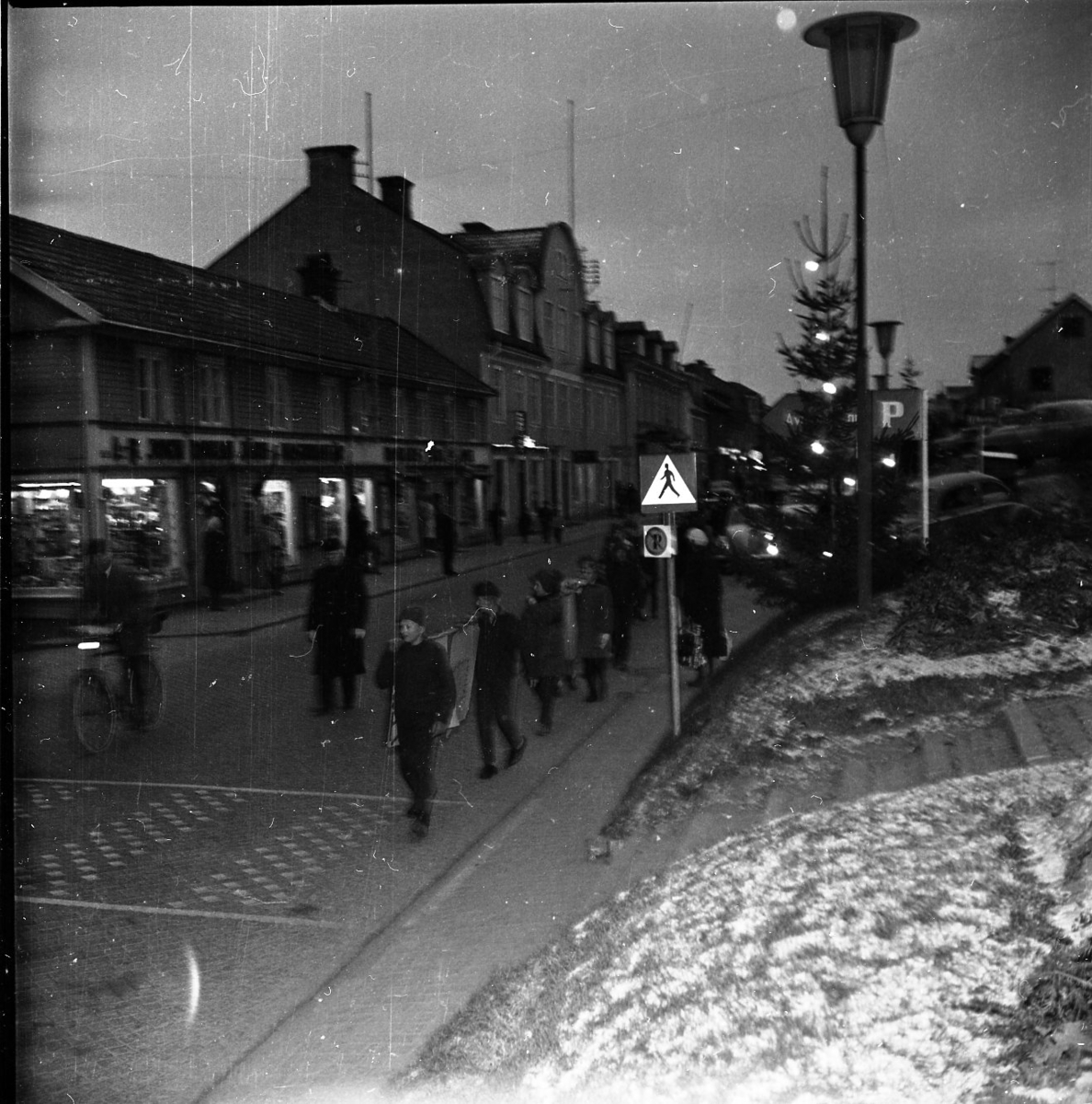 Ungdomar går i mörkret på Brahegatan. Tända julgranar på torgmuren till höger. Det ligger lite snö på marken.