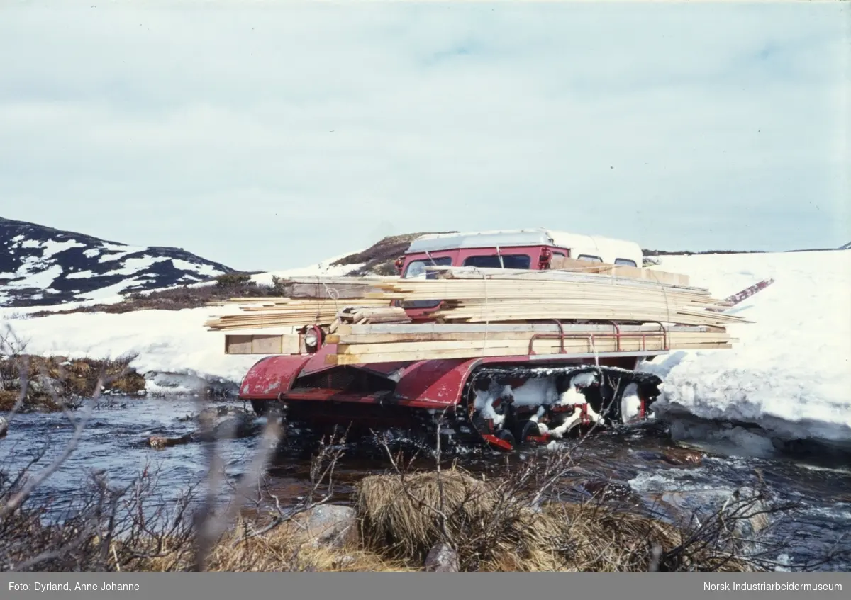 Materialfrakt av plank med beltebil over snø og elv i Grotvassdalen, Seljord