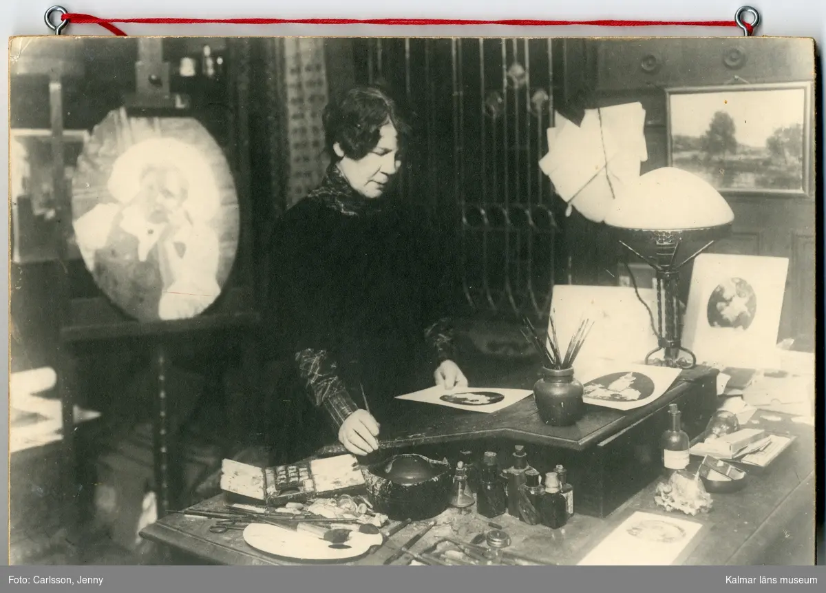 Svartvitt fotografi föreställande Jenny Nyström. Hon står vid ett bord i sin ateljé, hon har en pensel i ena handen och en teckning i den andra. Hon är svartklädd.