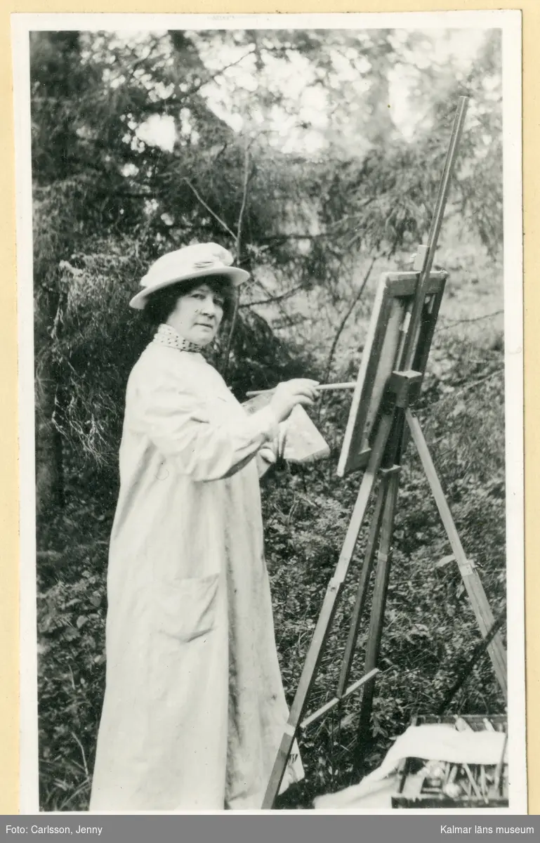 Jenny Nyström står vid ett staffli utomhus och målar. Hon är klädd i vit rock och vit hatt. Hon håller en palett i sin vänstra hand och en pensel i sin högra. Under staffliet ligger en låda med penslar m.m. på marken.