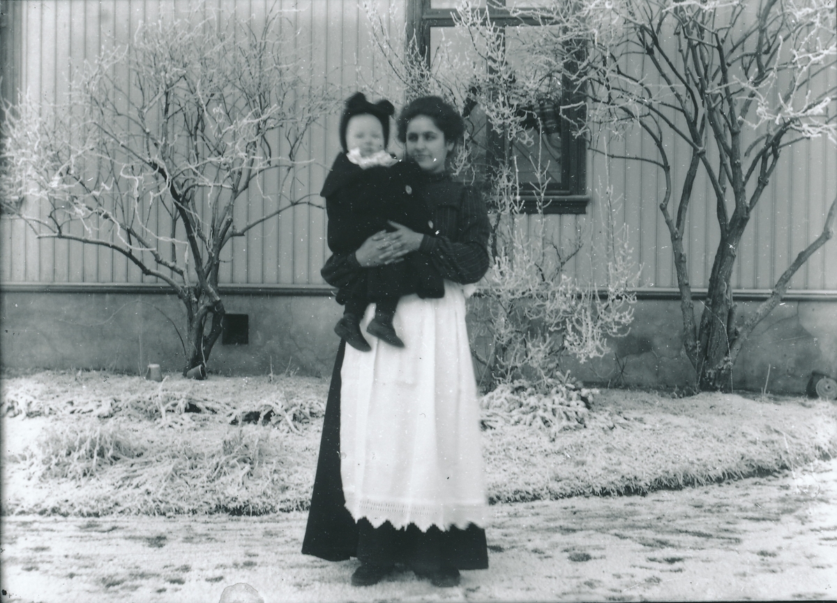 Ung kvinne holder lite barn på ca. 1 år i armene. Kvinnen er iført lang kjole med forkle med heklet kant nederst. De står utenfor bolighus i tre foran rimfrossen plen.