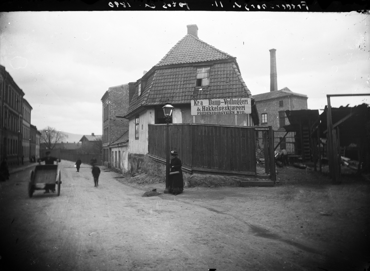 Gatebilde fra Fredensborgveien. Karer med håndtraller og kvinne som venter. Skilt til Kr. a. Damp-vedhuggeri & Hakkelseskjæreri.