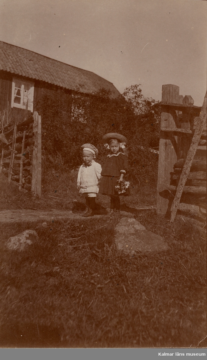Fotografi av två barn utomhus. En pojke Curt Nyström Stoopendaal till vänster? och en flicka till höger. Flickan är klädd i hatt, jacka, kjol, strumpor och skor. Barnen står mitt i en öppning i ett staket. I bakgrunden syns ett hus. I gräset till höger syns stora stenar.