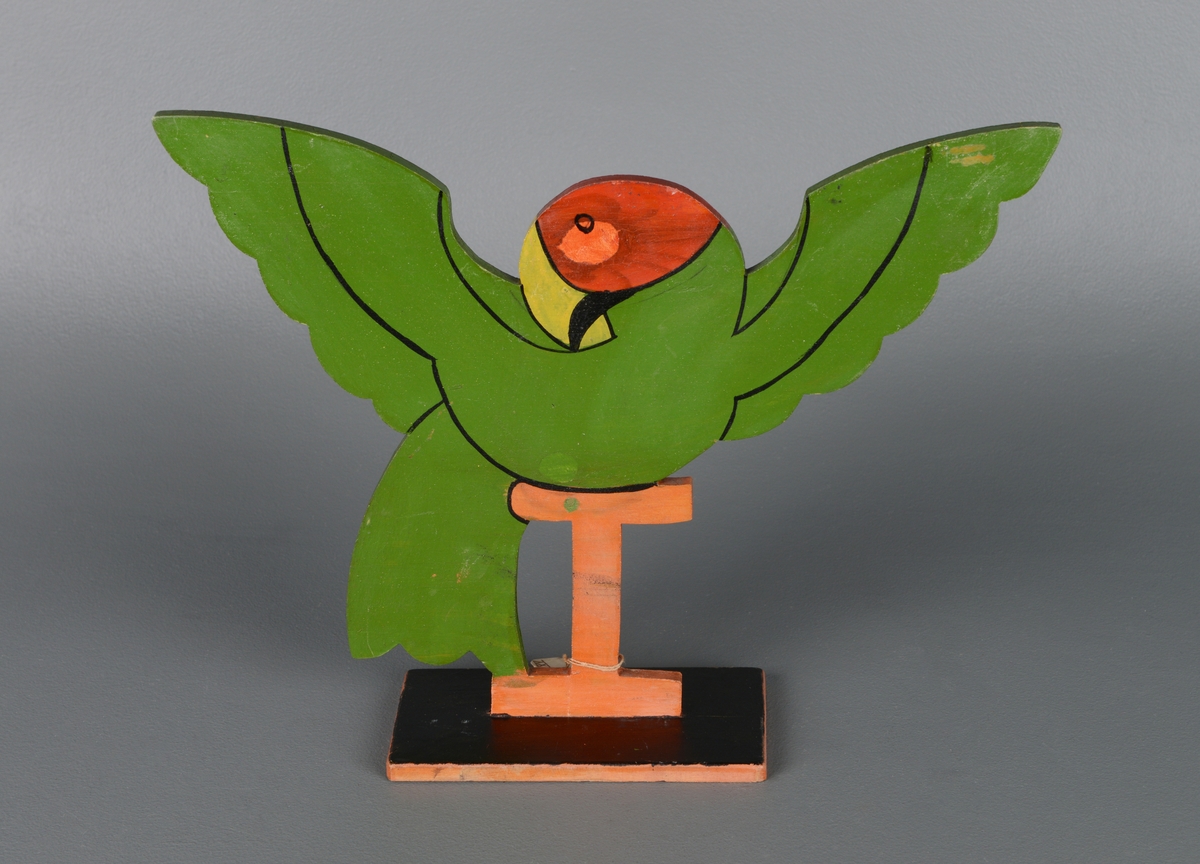 En stående stilisert figur av en papegøye på pidestall med vingene utslått. Figuren er laget av en tynn treplate montert på en rektangulær sortmalt treplate. Figuren er utskjært og malt på begge sider i rødt og grønt, med sorte linjer for øyne, vinger og nebb.