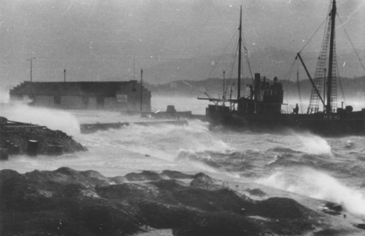 Motiv fra havnen i Ålesund under storm og springflo. En fiskebåt, M-9-HD(?), ligg ved Storneskaia.