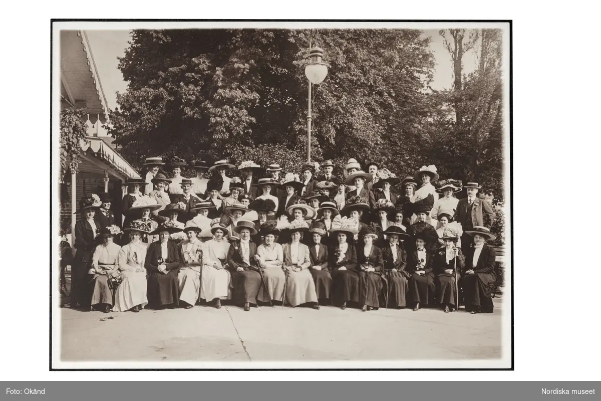 Ett grupporträtt av personalen på NK:s Franska damskrädderi på utflykt 1911. En grupp med sittande kvinnor i dräkt och hatt flankerade av några kostymklädda män. Den unge mannen längst bak, sjätte från vänster är Kurt Jacobsson, då nittonårig volontär hos Madame Suzanne.