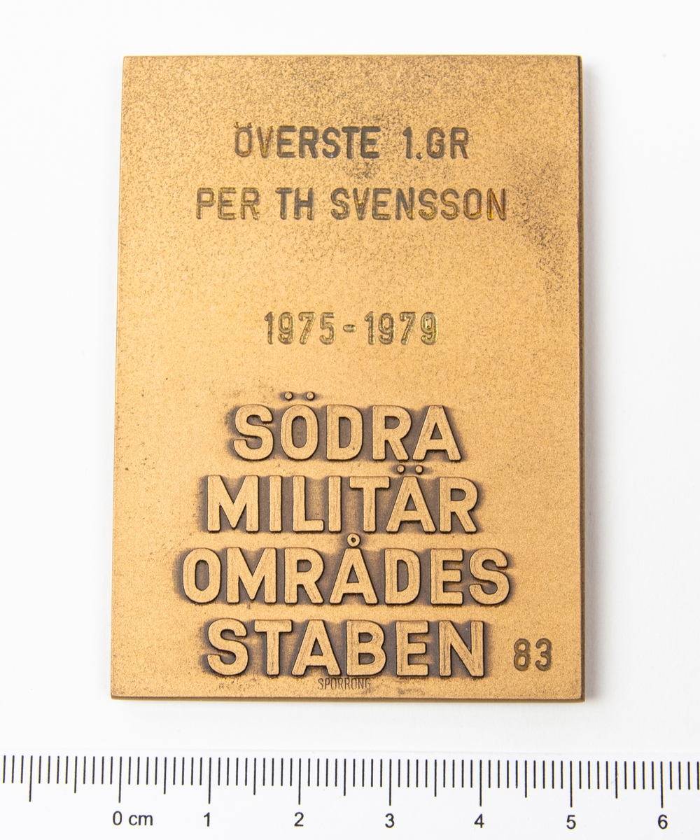 Plakett i ask. På framsidan ett svärd med ett S i relief. På baksidan står det: Överste 1 Gr Per Th Svensson 1975 - 1979 Södra Militär Områdes Staben 83.