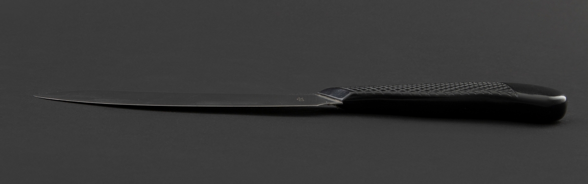 Skinkekniv i rustfritt stål og svart plastskaft med rutemønster. Skaftet er festet skrått til bladet. Spisst avsluttet blad.