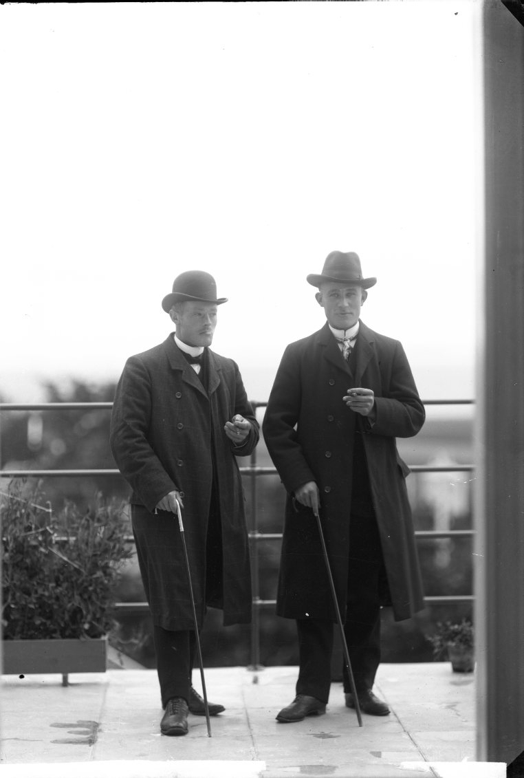 Två unga män med rock, hatt, spatserkäpp cigarett i händerna står på en balkong med utsikt mot Vättern. En av männen är Josef Gustafsson från Östanå.