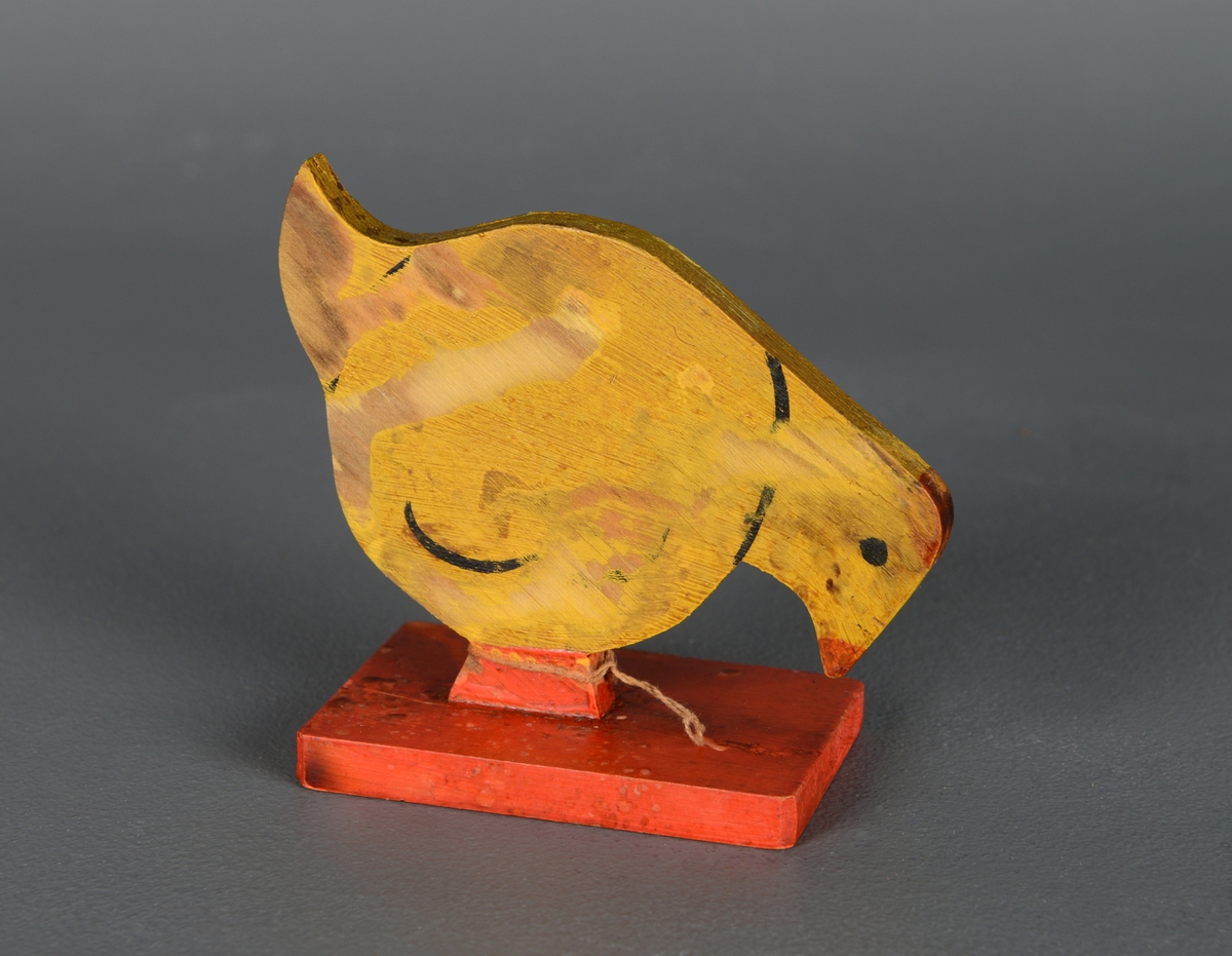 En liten, stående figur av en stilisert kylling laget av en tynn treplate montert på en rektangulær, rød treplate. Den er malt gul, og detaljene som vinger og øyne er markert i sort. Nebb og føtter er malt i rødt.
