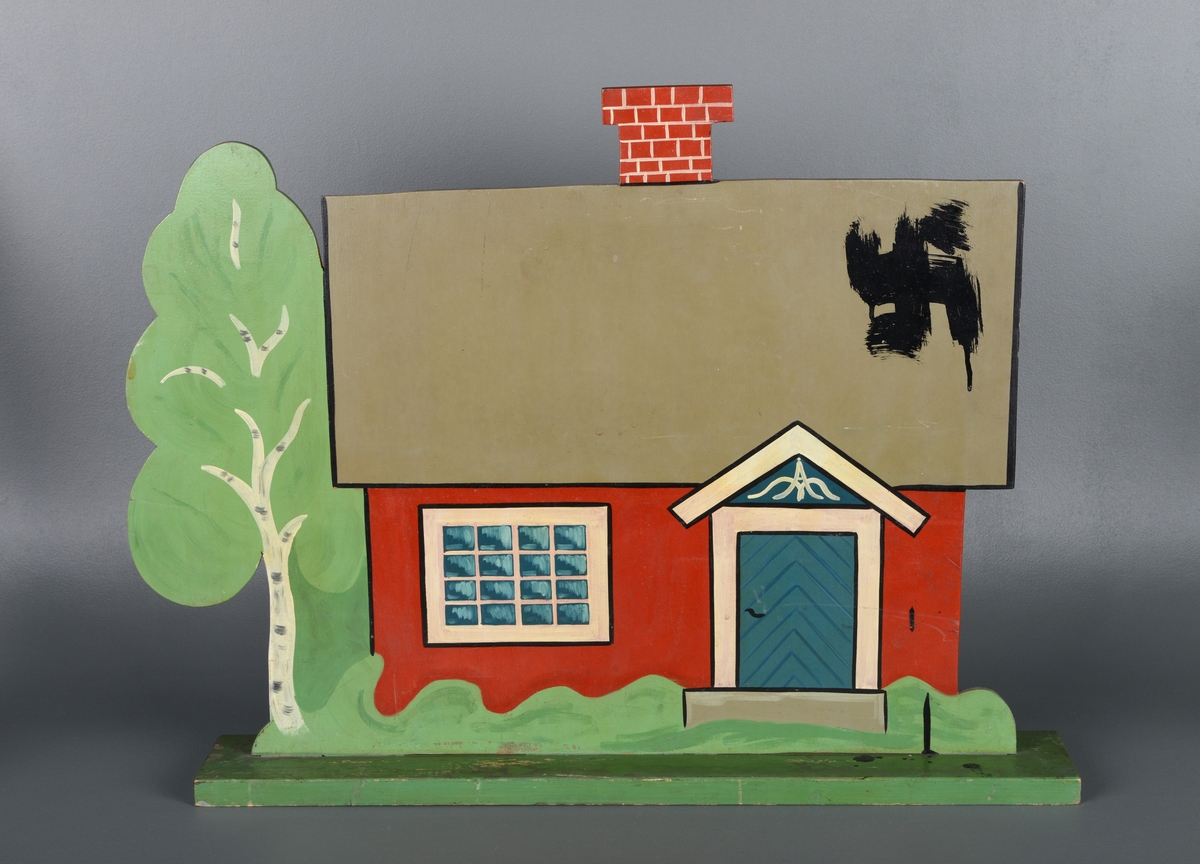 En stående figur av et hus med murpipe og et bjørketre laget av en tynn treplate montert på en rektangulær, grønn treplate. Øverst til høyre på taket er det malt et hakekors med store, sorte malestrøk. Sortmalingen er dryppet nedover huset og på platen. Huset er malt i rødt, med grått tak, blå dør og hvite vinduskarmer. På baksiden er figuren malt helt sort.