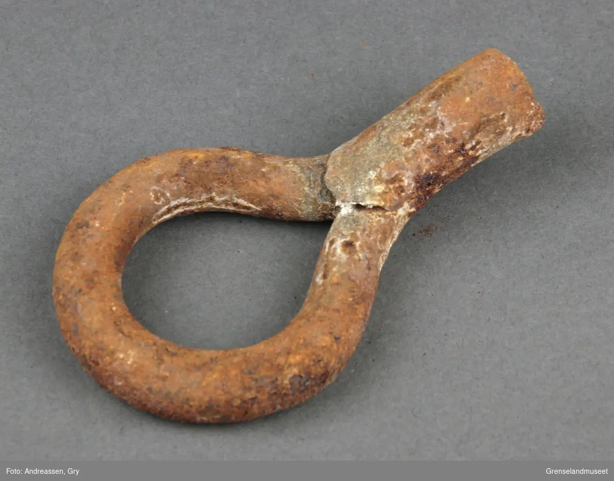 En øyebolt i jern, ringen er inntakt men selve bolten eller kroken er knukket av. Rusten.