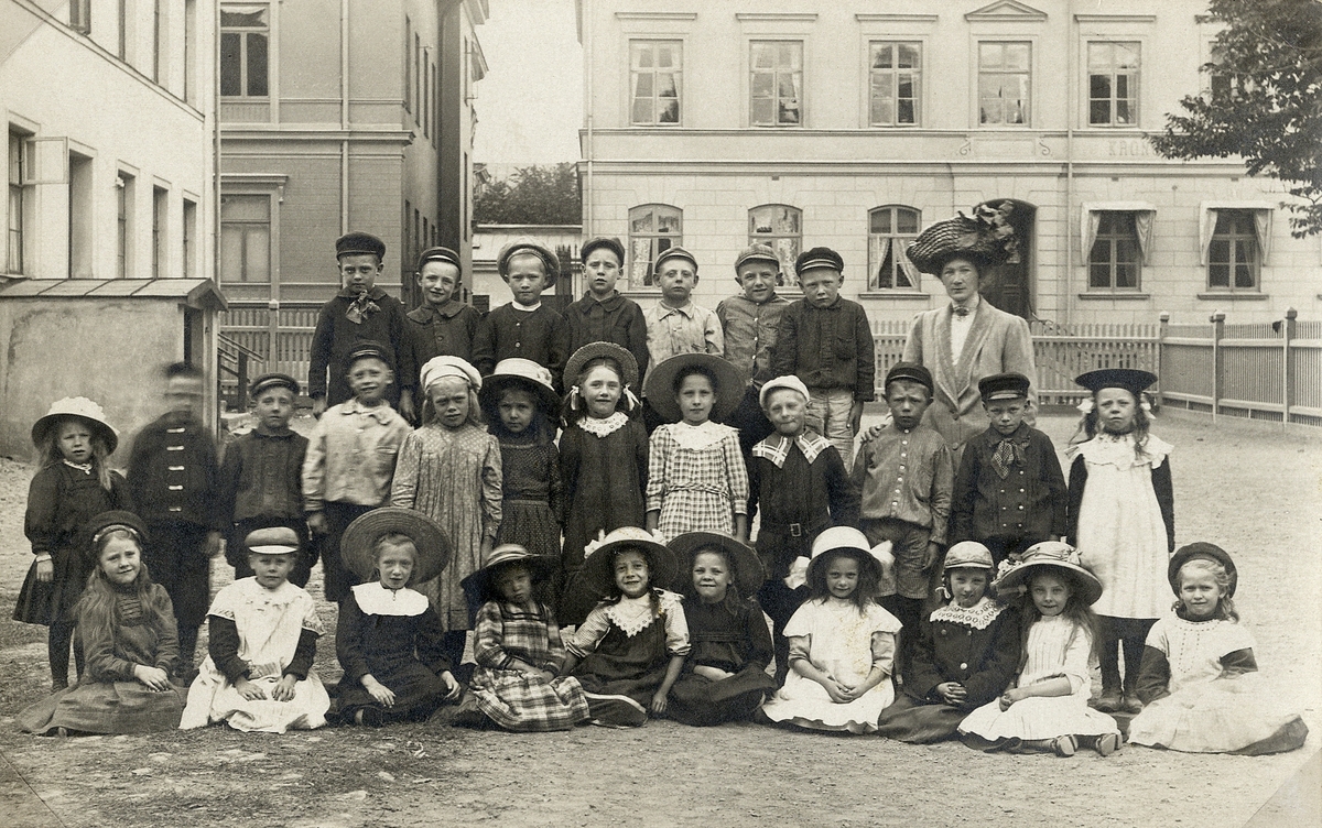 En skolklass har ställt upp sig för fotografen på skolgården till Sjögrenska skolan, Växjö, ca 1912. 
I bakgrunden syns bl a hörnet av Hypoteksbanken på Kungsgatan 12 och dåv. Kronobergs Sparbank på Nygatan 8.
Vykort.