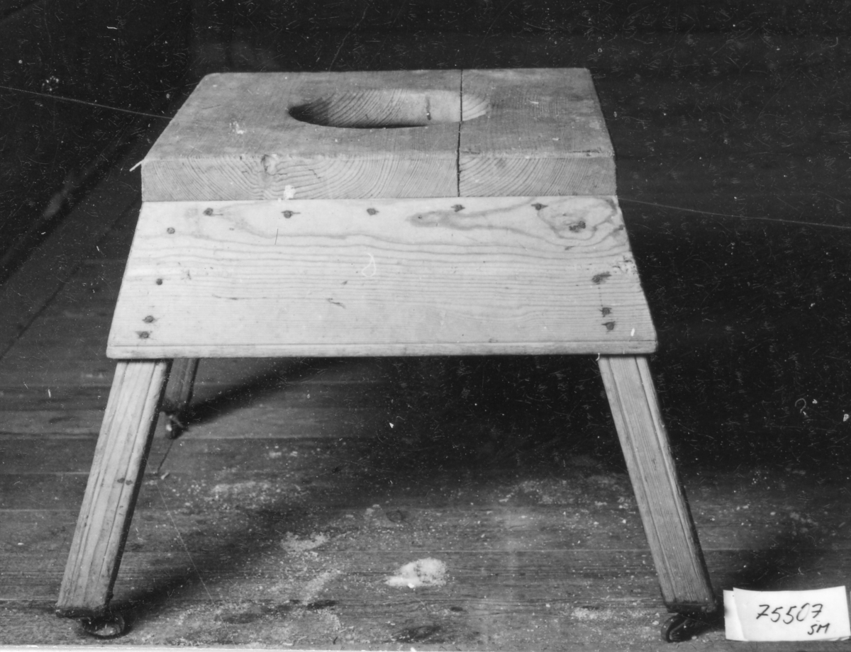 Omålad gåstol med kvadratisk skiva försedd med ett större hål i mitten. 4 stycken 4-sidiga utställda ben med hjul.