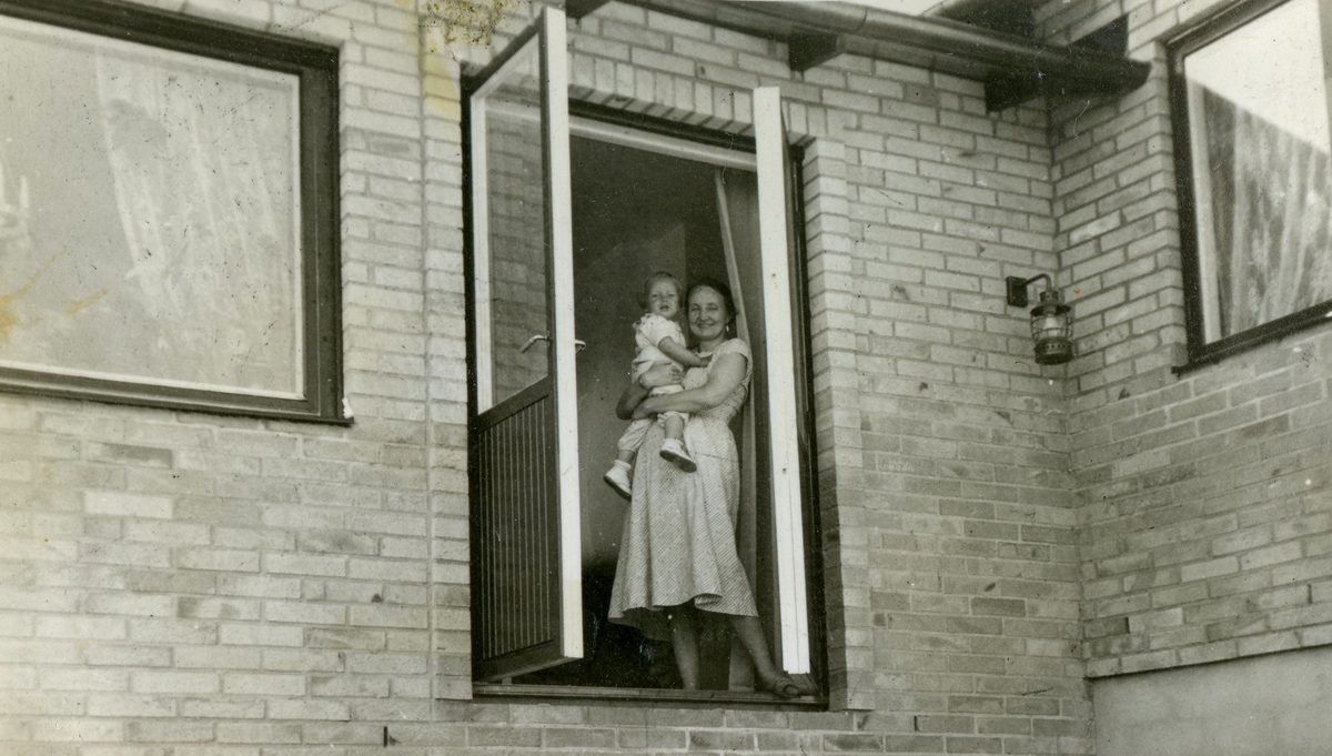 Mary Ekman håller sonen Manne (född 1955) i famnen, april 1957. De står i balkongöppningen till familjens nybyggda villa (då utan trappa och altan), Villa Maribo på Streteredsvägen 29.