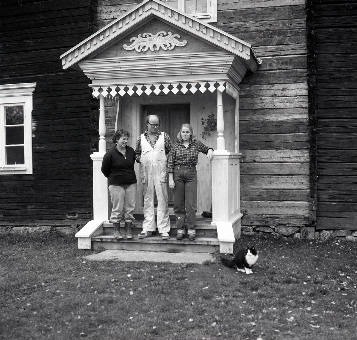 Tre personer står uppställda på en förstukvist för fotografering. TV-inspelning om brokvistar på Hymmersbacken i Alfta, 1981. Ett timrat hus renoveras.