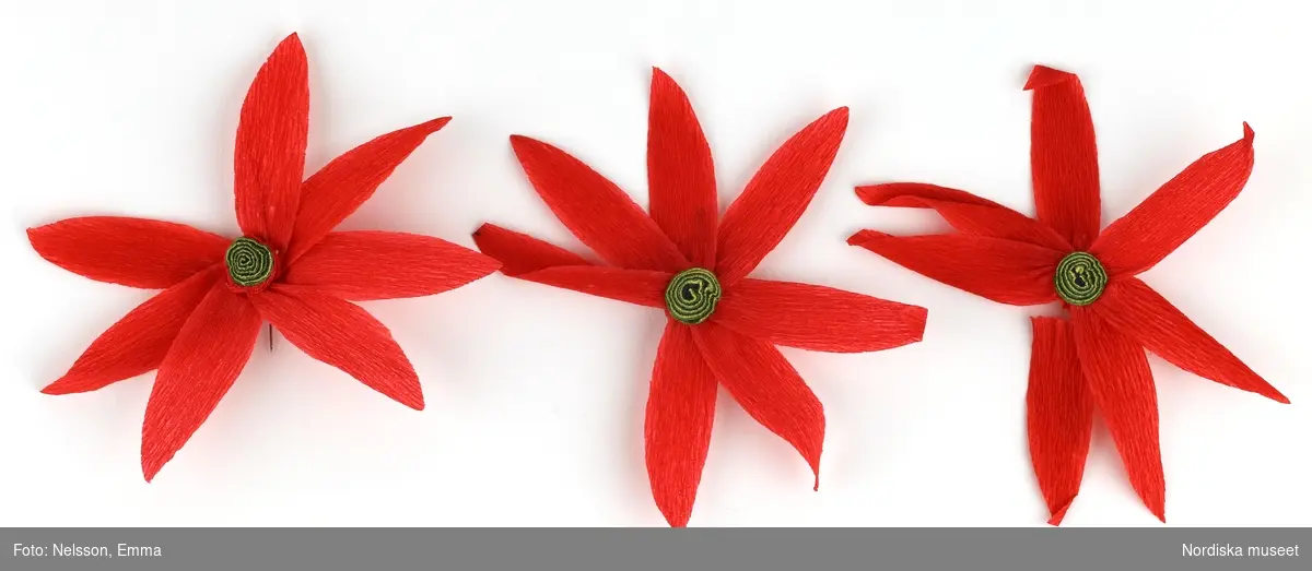 a-l) Tolv stycken hemtillverkade julgransprydnader av rött kräpp-papper i form av blommor/julstjärnor, med upphängningsanordning av knappnålar. 

Lena Kättström Höök 2019-03-21