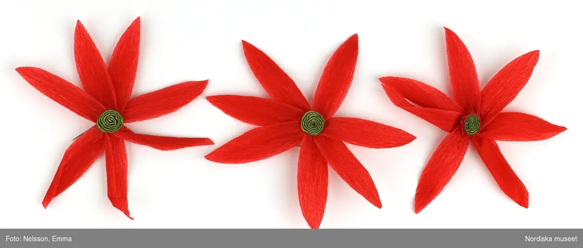 a-l) Tolv stycken hemtillverkade julgransprydnader av rött kräpp-papper i form av blommor/julstjärnor, med upphängningsanordning av knappnålar. 

Lena Kättström Höök 2019-03-21