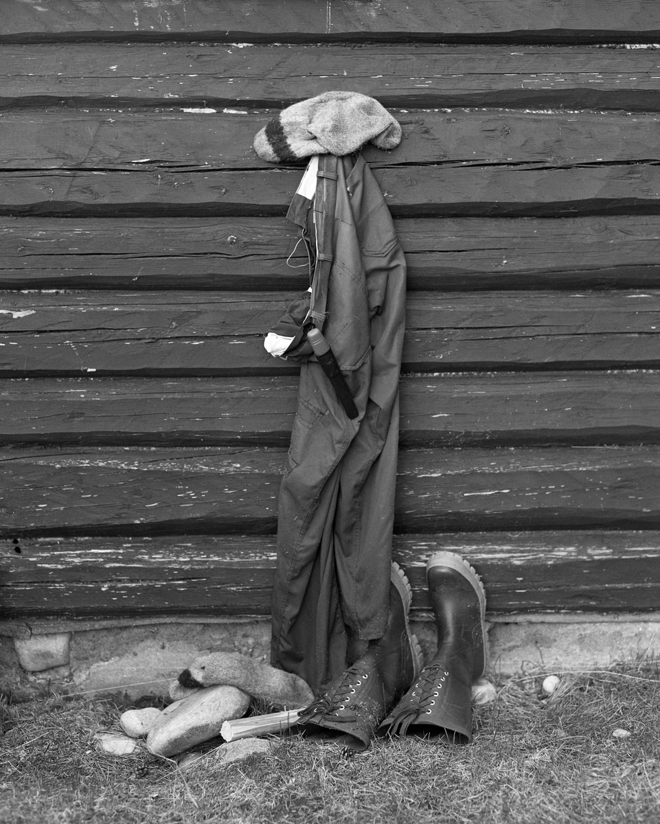 Arbeidsklær til tørk ved Stenbekkoia ved Feumdselva i Engerdal, fotografert våren 1986. Fotografiet er tatt mot en laftet koievegg, der det hang ei beavernylonbukse og en strikket ullsokk til tørk. Den andre sokken hadde falt ned på bakken, der den lå ved sida av de langskaftete gummistøvlene som lå på bakken med sålene mot husveggen. Skaftene lå en aning nedoverskrånende med en åpning som innebar et visst innsig av luft.