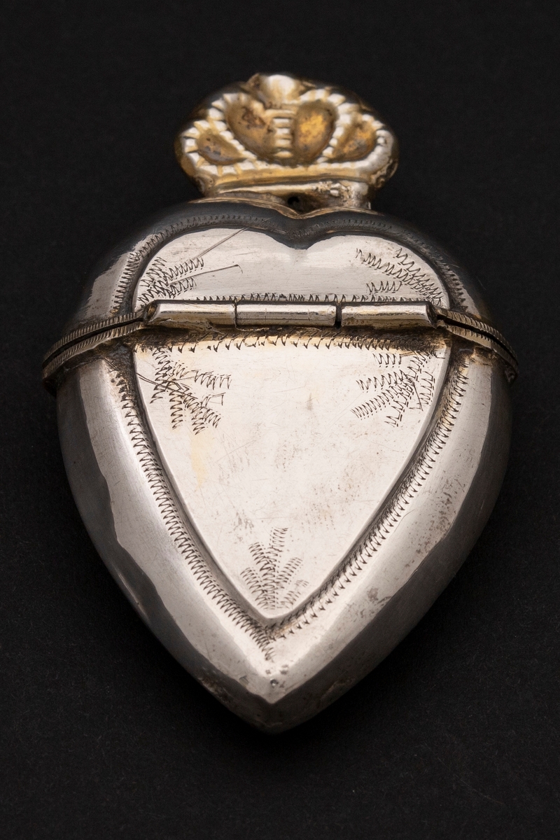 Hjerteformet luktevannshus i sølv med sammenklemt, forgylt krone på lokket. Dekorert med strekornamentikk; sikksakklinjer i border og satt sammen til bladlignende former. Innvendig forgylt. Tynt materiale.