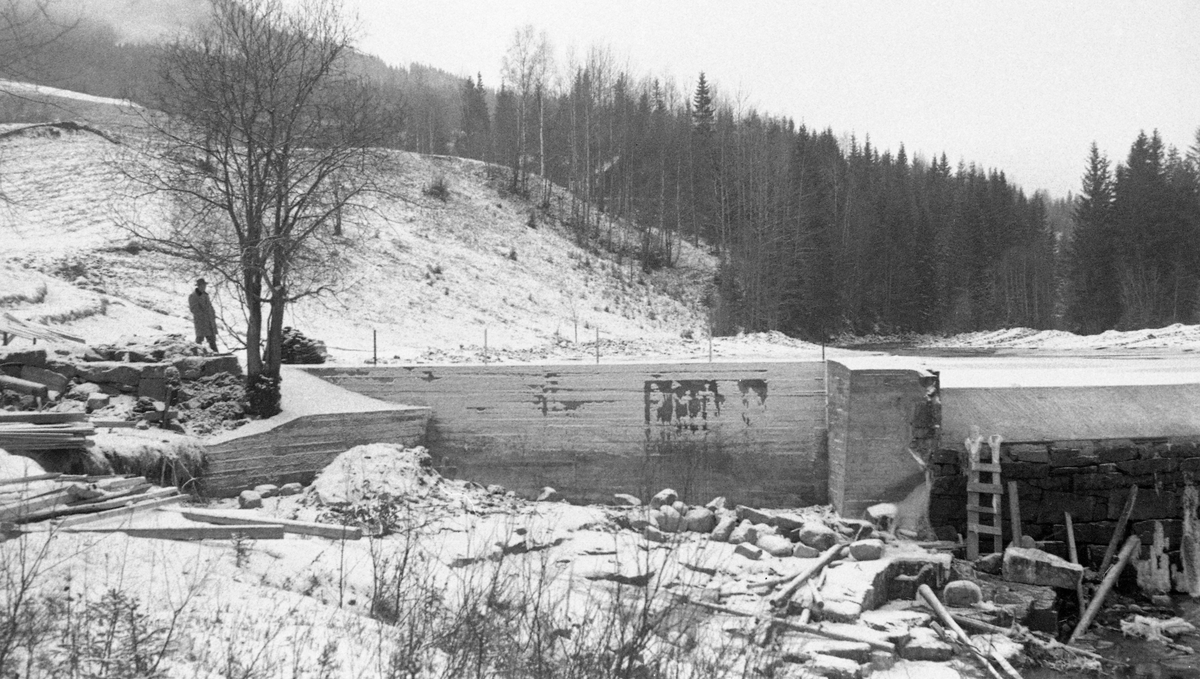 Mølledam, sannsynligvis ved Flaen mølle i Hurdalselva (Høverelva) i Akershus, fotografert etter en reparasjon i 1957. Bildet er tatt i motstrøms retning, mot en nystøpt damarm. Til høyre for den ser vi en del av flomløpet sentralt i vassdraget, der den ellers tørrmurete dammen var påstøpt en betongterskel, antakelig for å øke volumet i det bakenforliggende vannmagasinet. Fotografiet ble antakelig tatt en høstdag, like etter at den første snøen hadde lagt seg på grasvollene ned mot dammen. En mann med hatt, antakelig en av funksjonærene fra Glomma fellesfløtingsforening, sto og betraktet den nye damarmen.