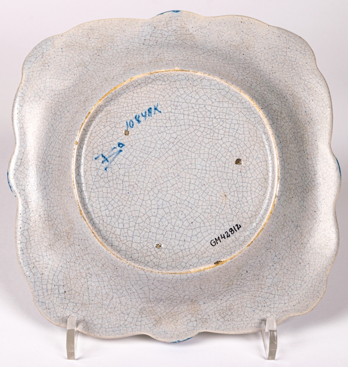 Fyrkantig assiett i lergods från Bo Fajans, modell 1084, formgiven av konstnär Allan Ebeling 1925. Glasyr AK: gråblåtonad antik konstglasyr (krackelerad) med handmålad dekor i blått i spegeln och på brämen.