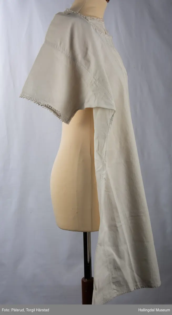 Hvit nattkjole eller serk i bomull med heklet kant rundt ermer og hals. Et hull er bøtt med en firkantet, hvit stoffbit midt framme nederst.