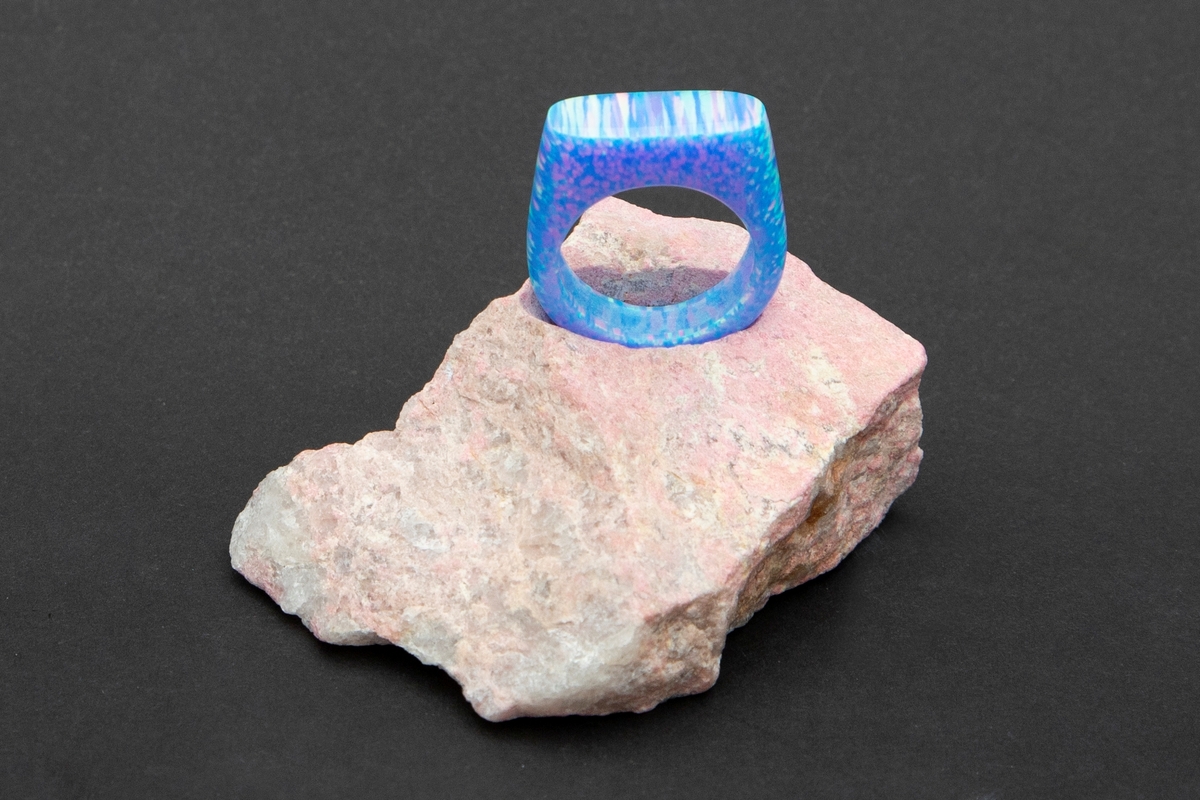 Lys blå ring med regnbue eller glittereffekter på rosa steinpodium. Ringen er lagd av syntetsik opal (bello) og podieet er av thulitt.