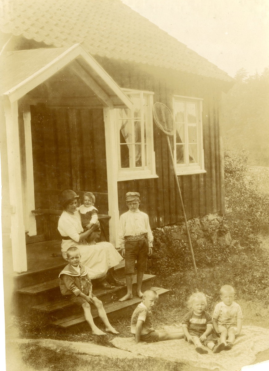 Kvinne og seks barn på trappen til lite hus. Hov på veggen. 1900 -20 ca.