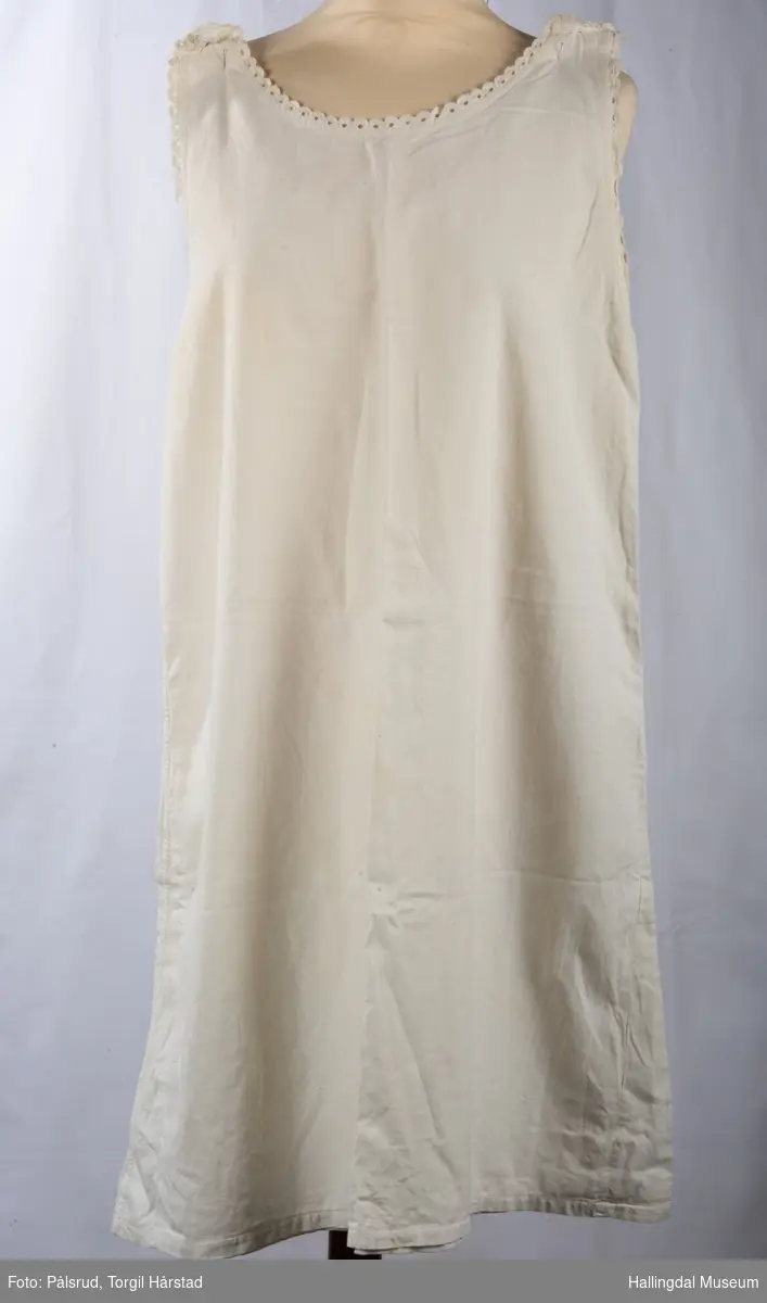 Hvit nattkjole i bomull med hvite blonder rundt hals og ermer. En hvit og en grå metallknapp på hver sin skulderstropp holder kjolen oppe.