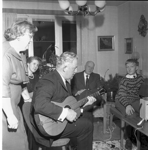 En kvinna står och sjunger medan en man spelar på en gitarr. Runt honom sitter åhörare och lyssnar.