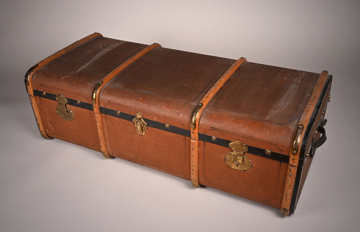 Koffert laget av tre, papp og skinn. Innsiden er kledd med papir. Den lukkes i front med tre beslag. På sidene er det festet bærehåndtak av skinn.