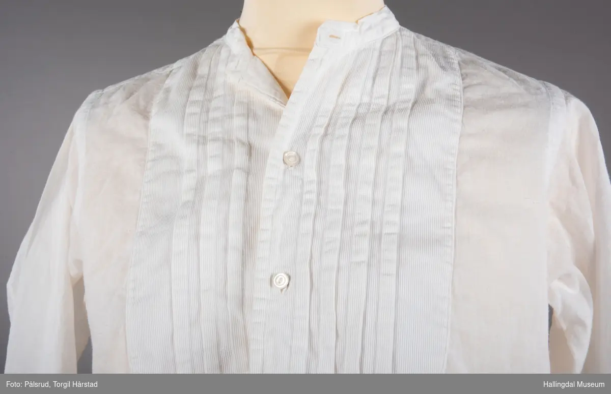 En hvit herreskjorte i bomull med folder på skjortebryst og to knapper. Uten krage i hals, med to knapphull i halslinningen og et knapphull bak. To knapphull i hver ermelinning til mansjettknapper. Splitt i sidene.